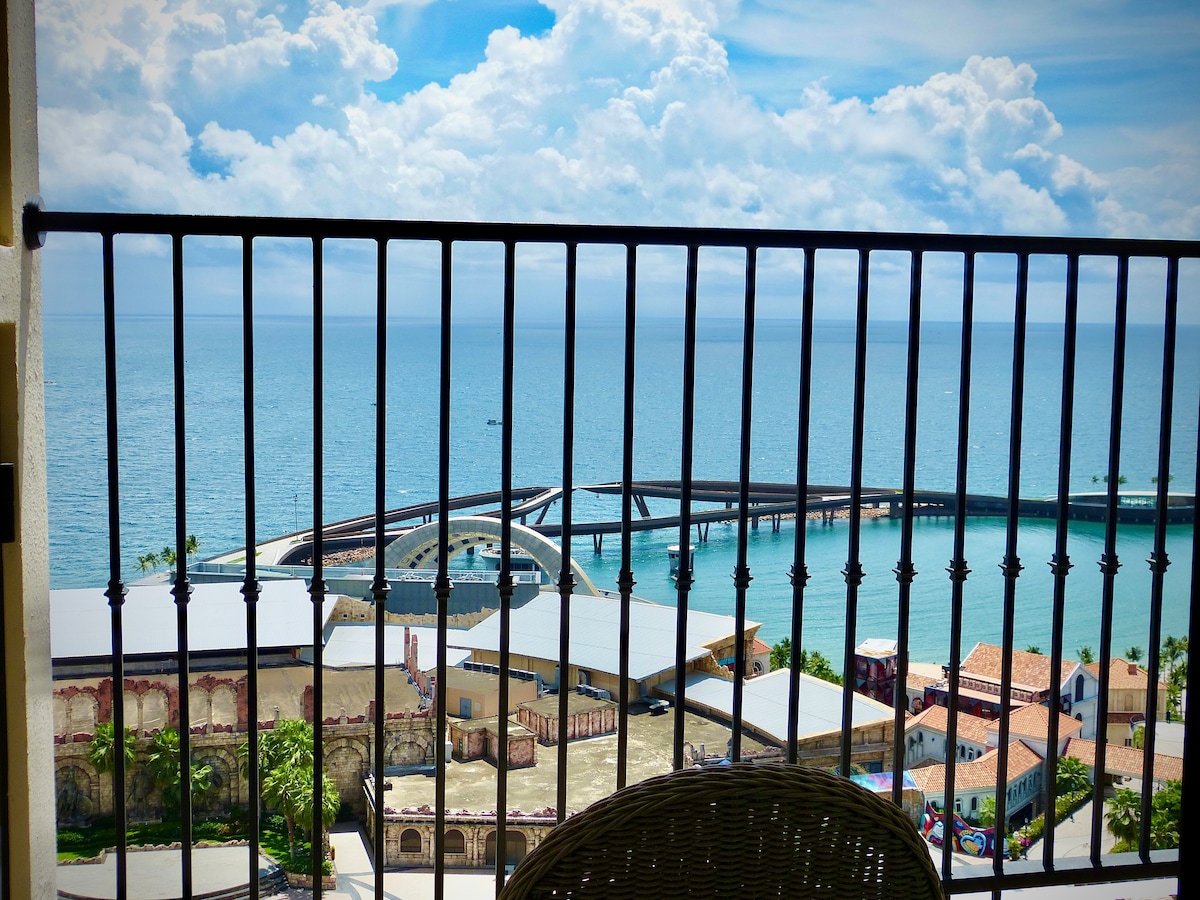 A scenic view from a balcony is captured, showcasing the vast ocean and a shoreline with vibrant blue waters. A structure can be seen in the foreground, with a curved pathway leading into the water. Fluffy clouds fill the sky, enhancing the serene atmosphere.