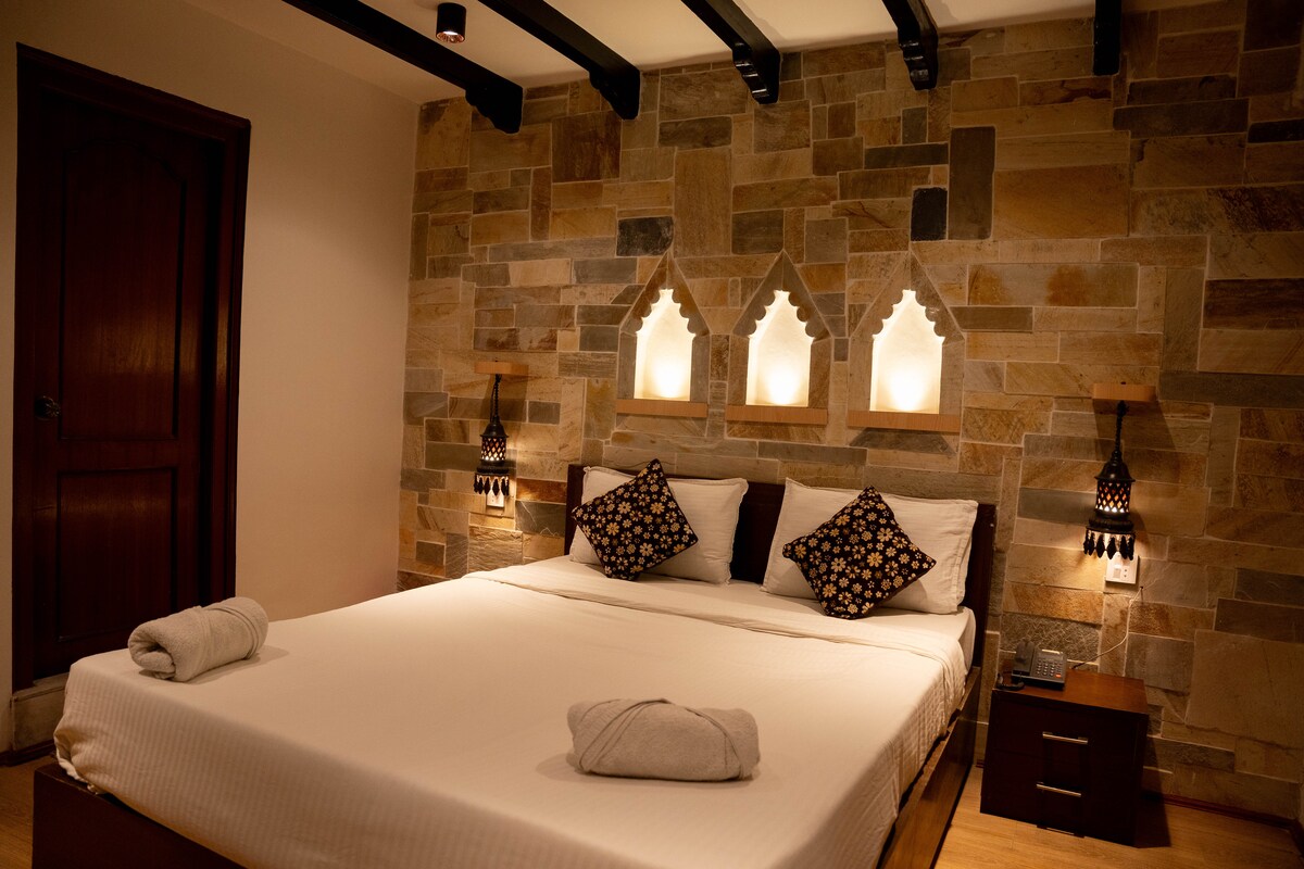 A cozy bedroom features a spacious bed adorned with patterned pillows and neatly rolled towels. The room's stone wall is complemented by soft lighting from decorative sconces. An intricately designed window cutout adds character, while wooden details enhance the serene ambiance.