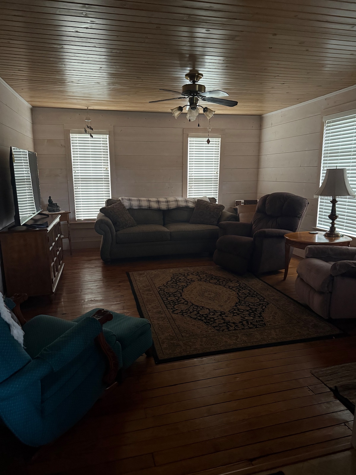 A cozy living area features a mix of comfortable seating, including a plush sofa and a reclining chair. Natural light filters through two window treatments, while a ceiling fan adds a rustic charm. A patterned area rug enhances the warm wooden flooring.