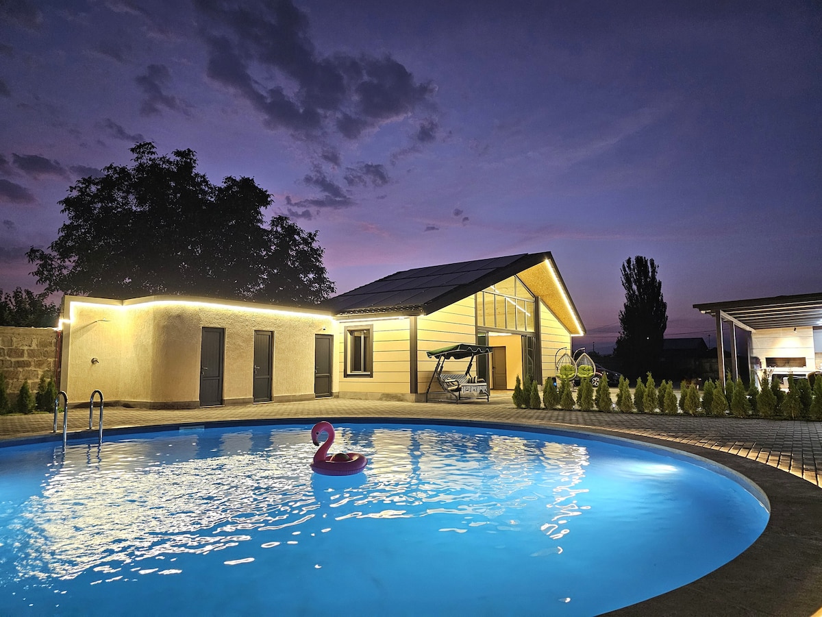 A modern rest house is situated beside a large swimming pool, illuminated by soft lighting. A colorful flamingo float is visible in the water. The structure features a bright exterior with large windows, and a covered seating area can be seen in the background.