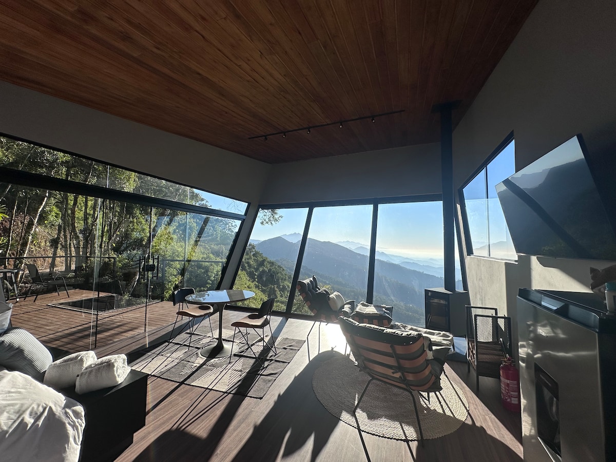 A modern living area features large glass windows offering panoramic mountain views. A wood ceiling contrasts with sleek furniture, including a round table and lounge chairs positioned thoughtfully. A cozy seating area is visible, inviting relaxation beside the fireplace, with natural light flooding the space.