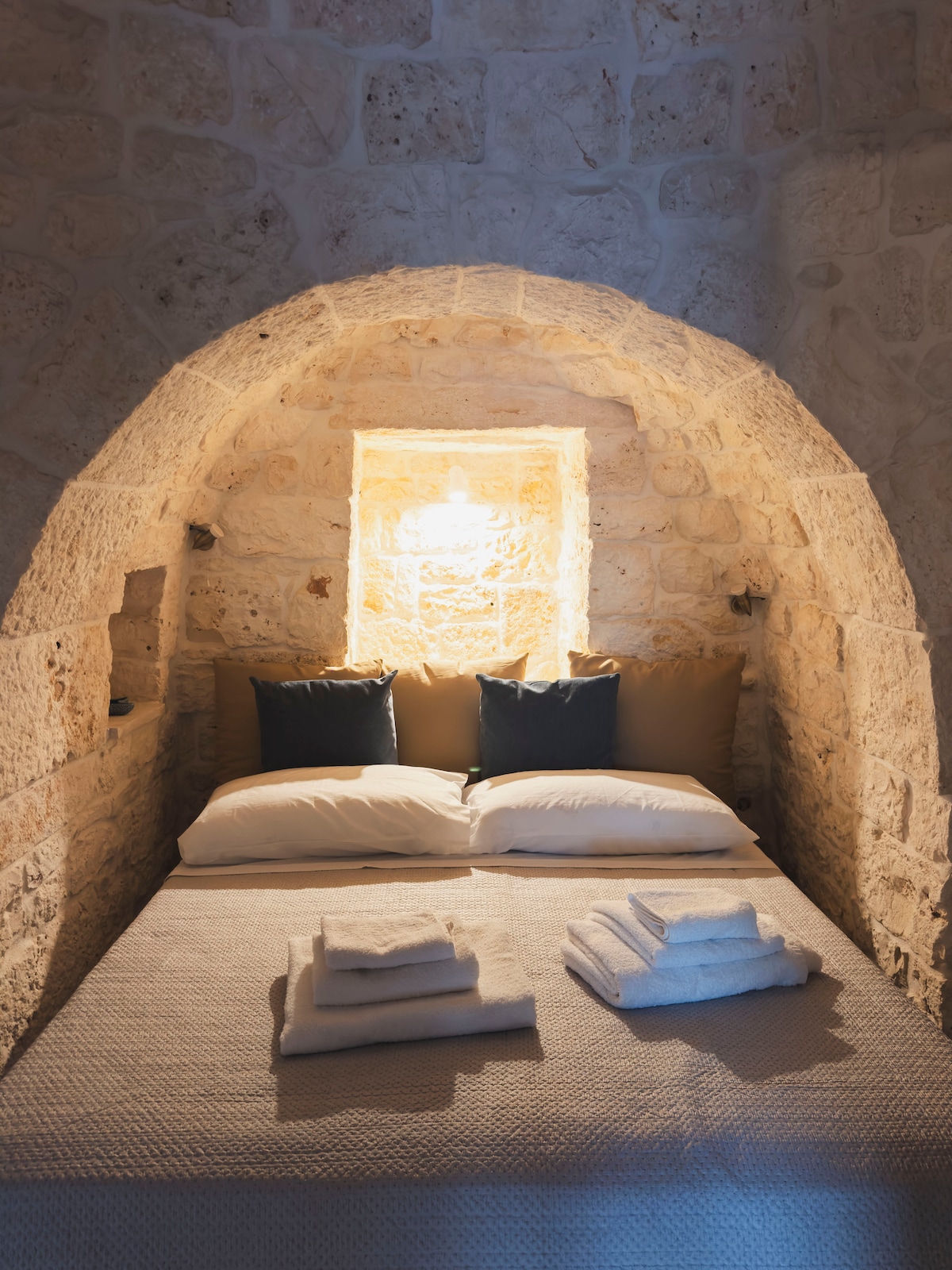 A cozy sleeping nook features a spacious bed adorned with soft pillows and neatly folded towels. The stone wall creates a rustic ambiance, while a warm light fixture above the bed provides gentle illumination, highlighting the inviting textures of the bedding.