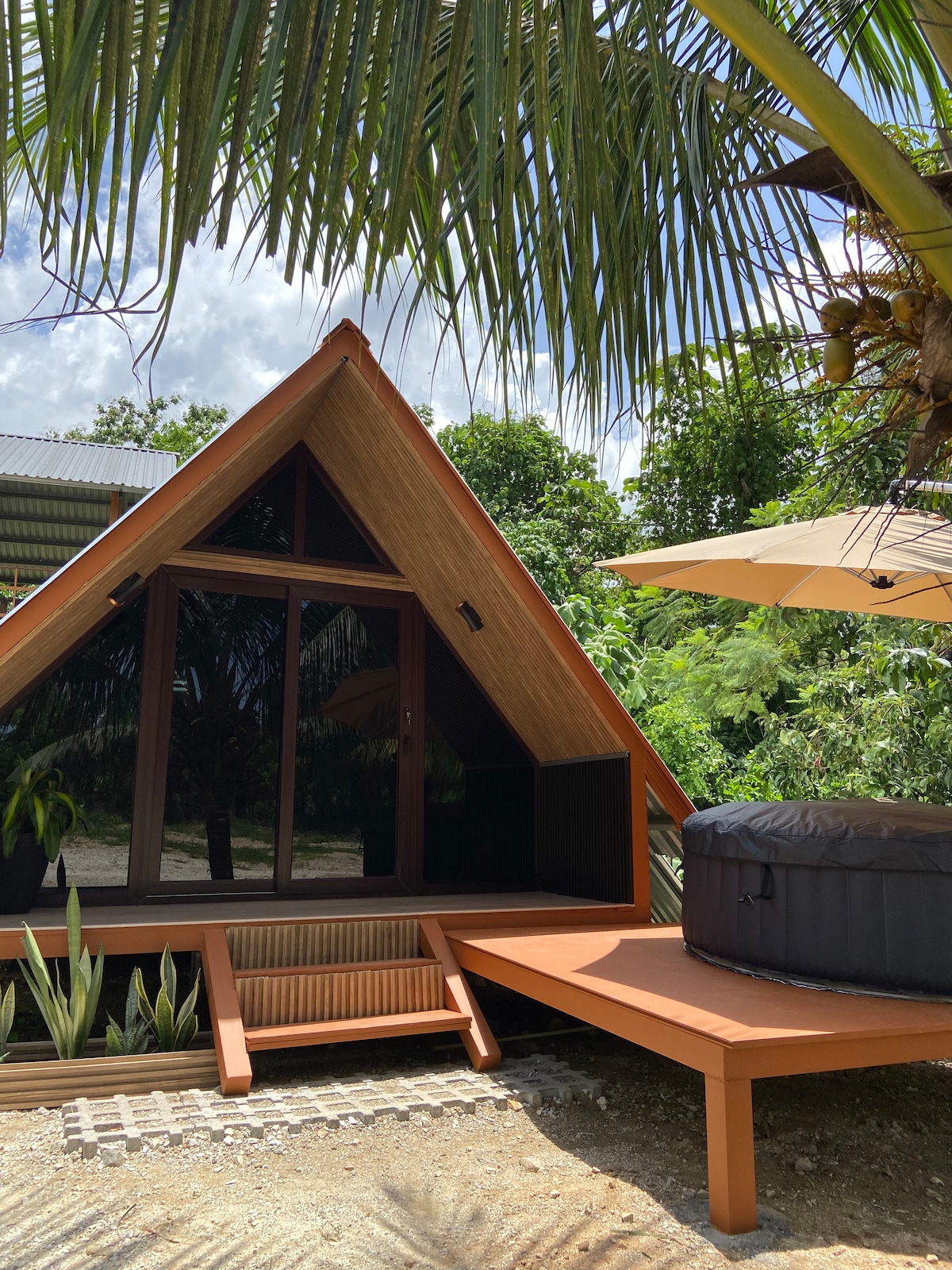 The exterior of a triangular-shaped cabin is showcased, featuring large glass windows that reflect the surrounding greenery. A sun deck extends toward a round hot tub, embraced by tropical plants and palm trees. An umbrella provides shade over the relaxation area.