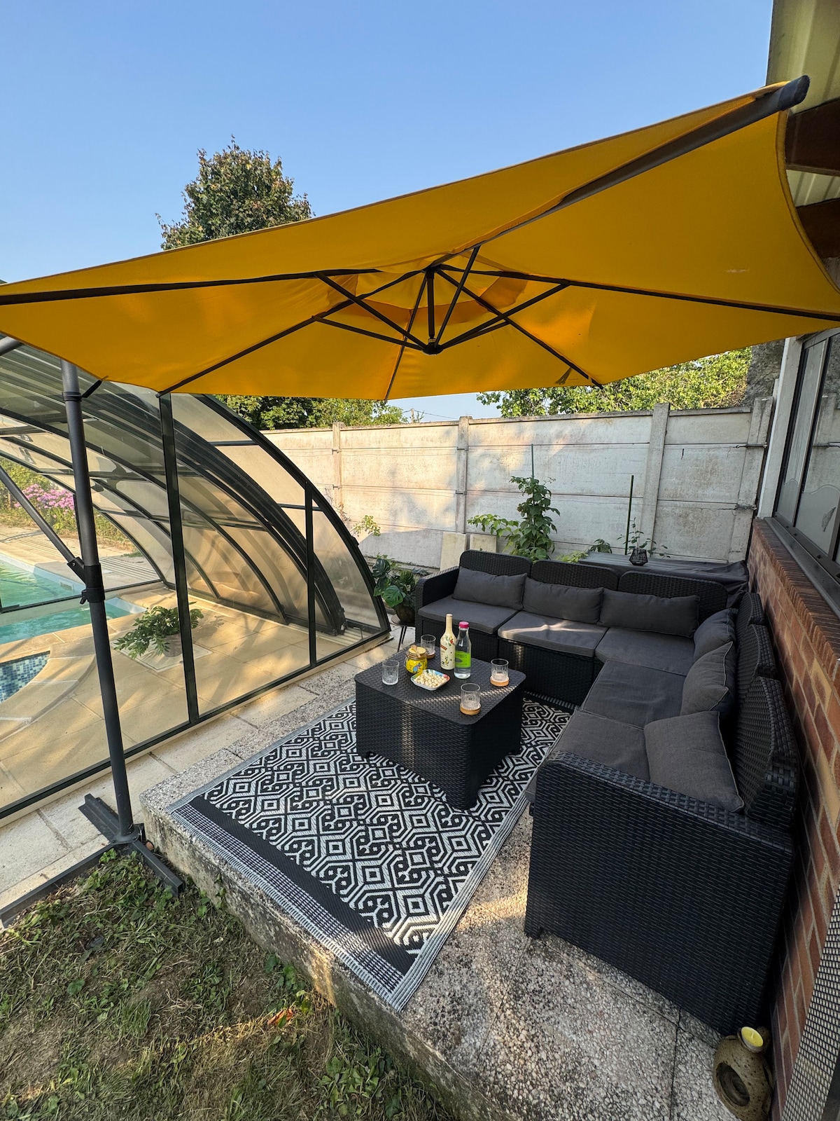 A shaded outdoor seating area is presented, featuring a dark wicker sectional sofa and a low table. An outdoor carpet adds texture, while a bright yellow canopy provides protection from the sun. The pool is partially visible in the background, enhancing the serene environment.