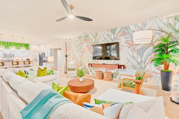 Amplio salón de la villa familiar de Orlando con una pared con hojas tropicales, un gran sofá modular blanco, una televisión inteligente, una silla de ratán, un ventilador de techo y exuberantes plantas en macetas