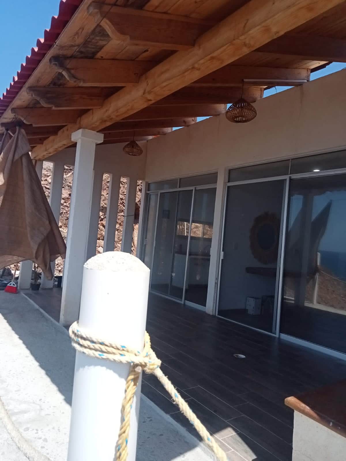 An outdoor terrace is showcased with wooden beams overhead and light fixtures hanging from the ceiling. Large glass sliding doors provide a view of the interior space, which includes light-colored walls and a mirror. Coastal scenery is visible in the distance.