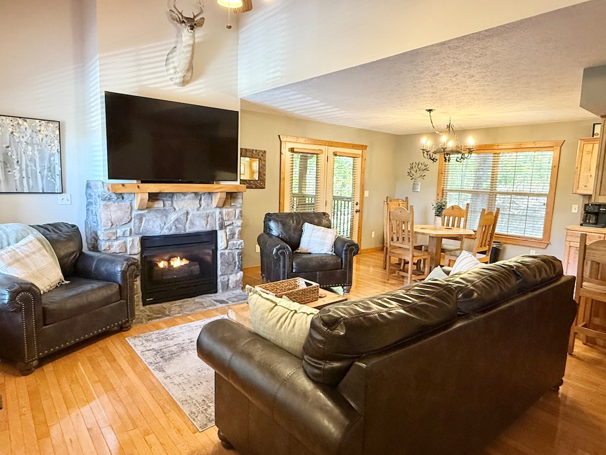A spacious living area features a stone fireplace beneath a large flat-screen TV. Comfortable leather sofas are arranged around a center coffee table. A wooden dining table with chairs is positioned nearby, illuminated by a chandelier. Natural light enters through multiple windows.