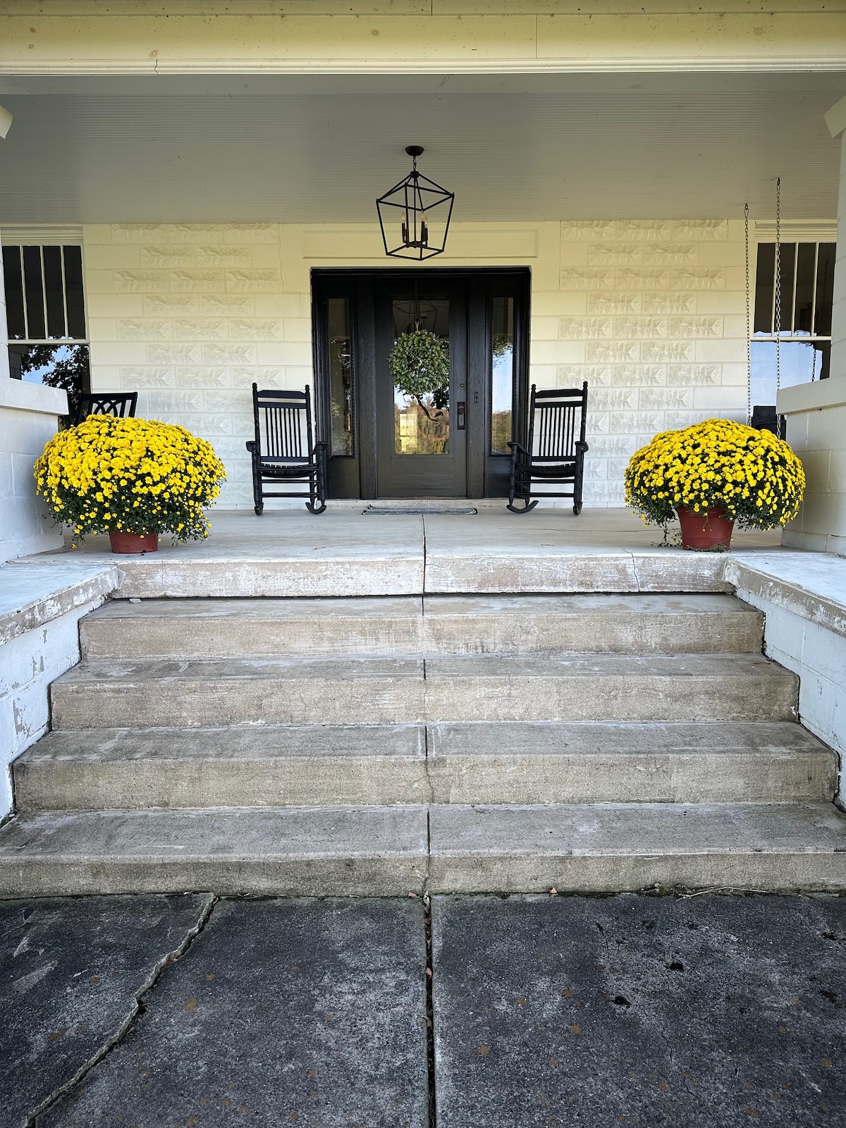 A welcoming entrance is depicted with two black rocking chairs positioned beside yellow chrysanthemum planters. The concrete steps lead up to a large double door, framed by a decorative lantern and a seasonal wreath, enhancing the charm of the farmhouse exterior.