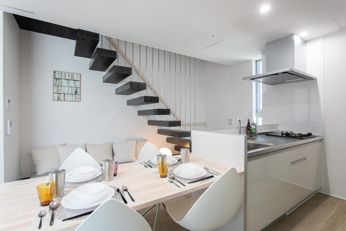 A modern dining area is presented, featuring a light wooden table set with plates and glasses. A cozy seating area with cushions is seen in the background. The open kitchen, equipped with sleek appliances, showcases a minimalist design. A stylish staircase leads to an upper level.