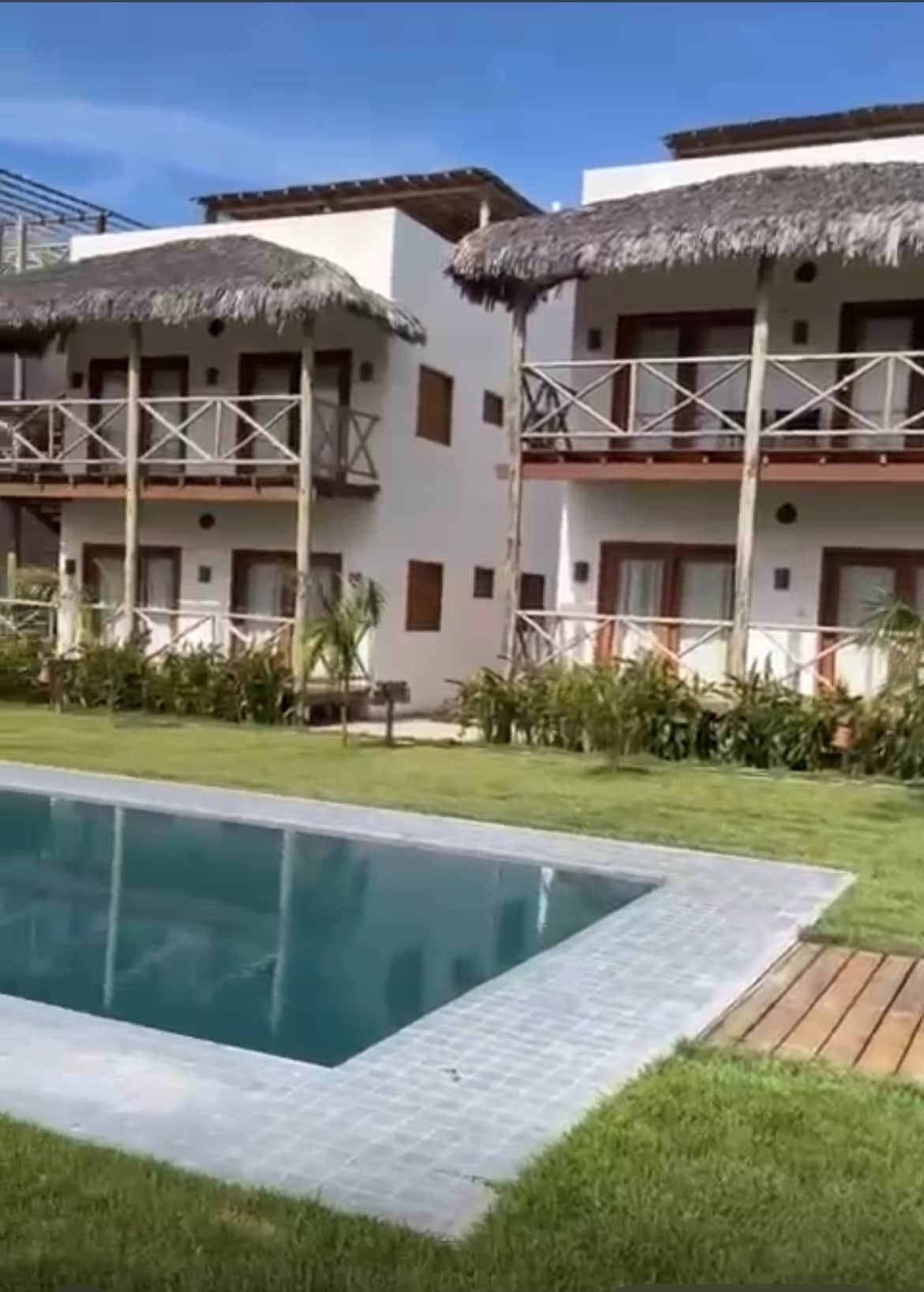 A tranquil pool is surrounded by well-maintained grass and lush greenery. Two multi-story buildings with thatched roofs feature balconies, providing a welcoming view of the outdoor area. The layout suggests a shared space, ideal for relaxation and gathering.