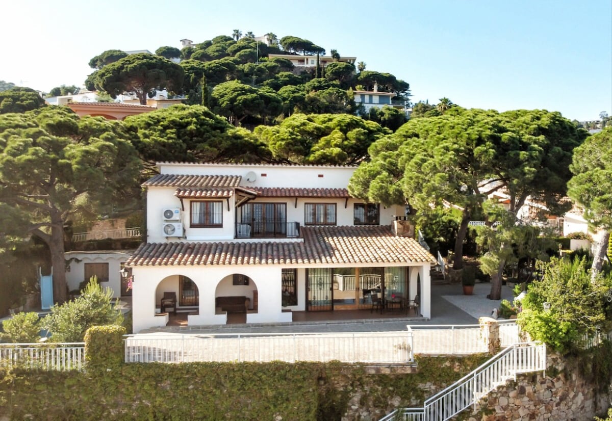 The Mediterranean-style villa is displayed from a frontal view, showcasing its charming architecture. A spacious terrace is visible, lined with potted plants. The villa is surrounded by lush, century-old pine trees, creating a serene backdrop in this gated urbanization.