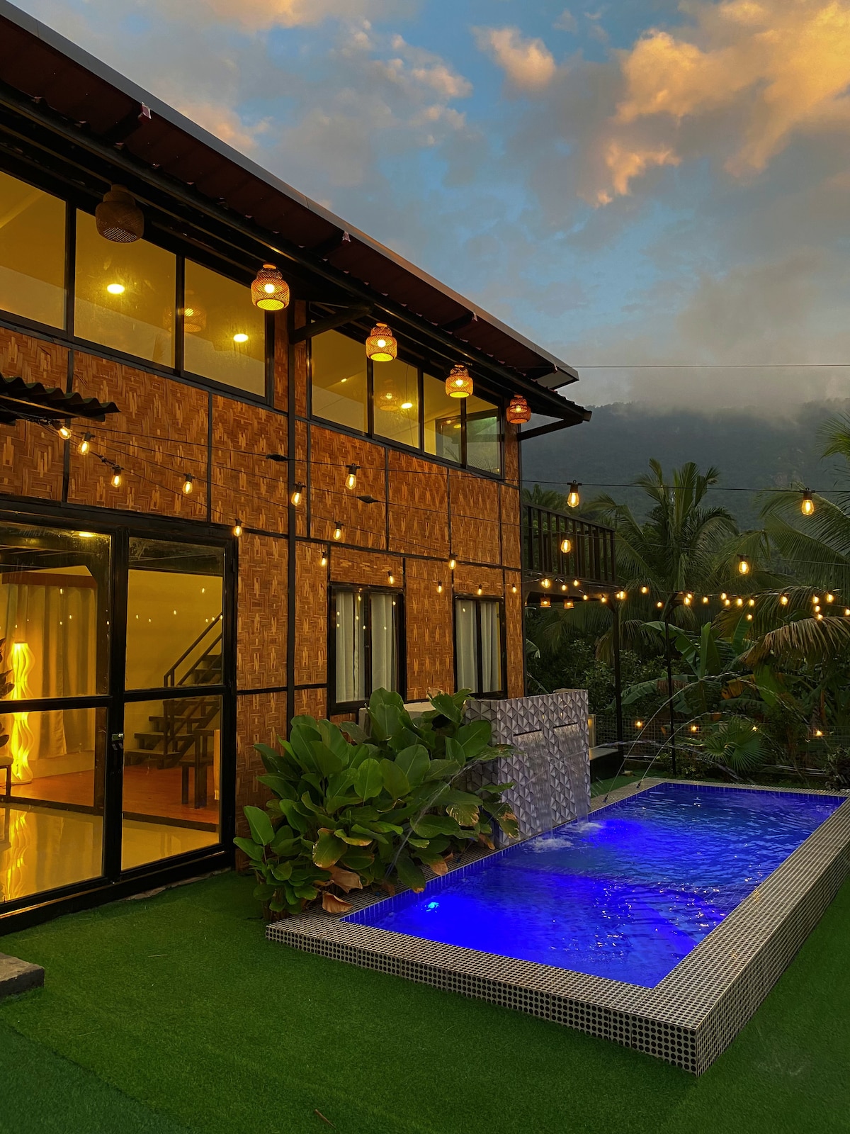 A two-story villa is illuminated by warm lights, showcasing a small swimming pool with vibrant blue water. Lush plants are positioned alongside the pool, and a serene mountain backdrop is visible behind the structure, enhancing the tranquil atmosphere of the outdoor space.