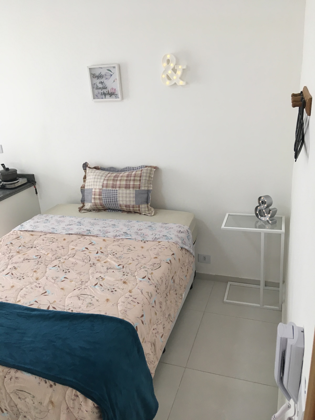 A bright and airy bedroom features a cozy bed with a patterned quilt and decorative pillows. A small side table is positioned beside the bed, displaying a sleek lamp. Light-colored walls and tiled flooring enhance the room's open feel.