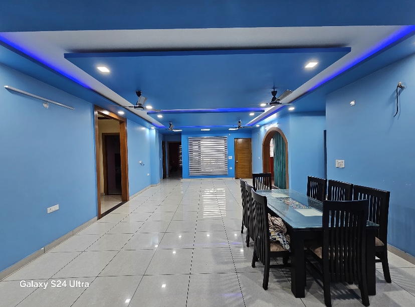 An elegant dining area features a long table surrounded by black chairs. Soft blue walls create a calming ambiance, with lighting set along the ceiling. A spacious living area is visible in the background, complemented by a sliding door with horizontal slats, inviting natural light.