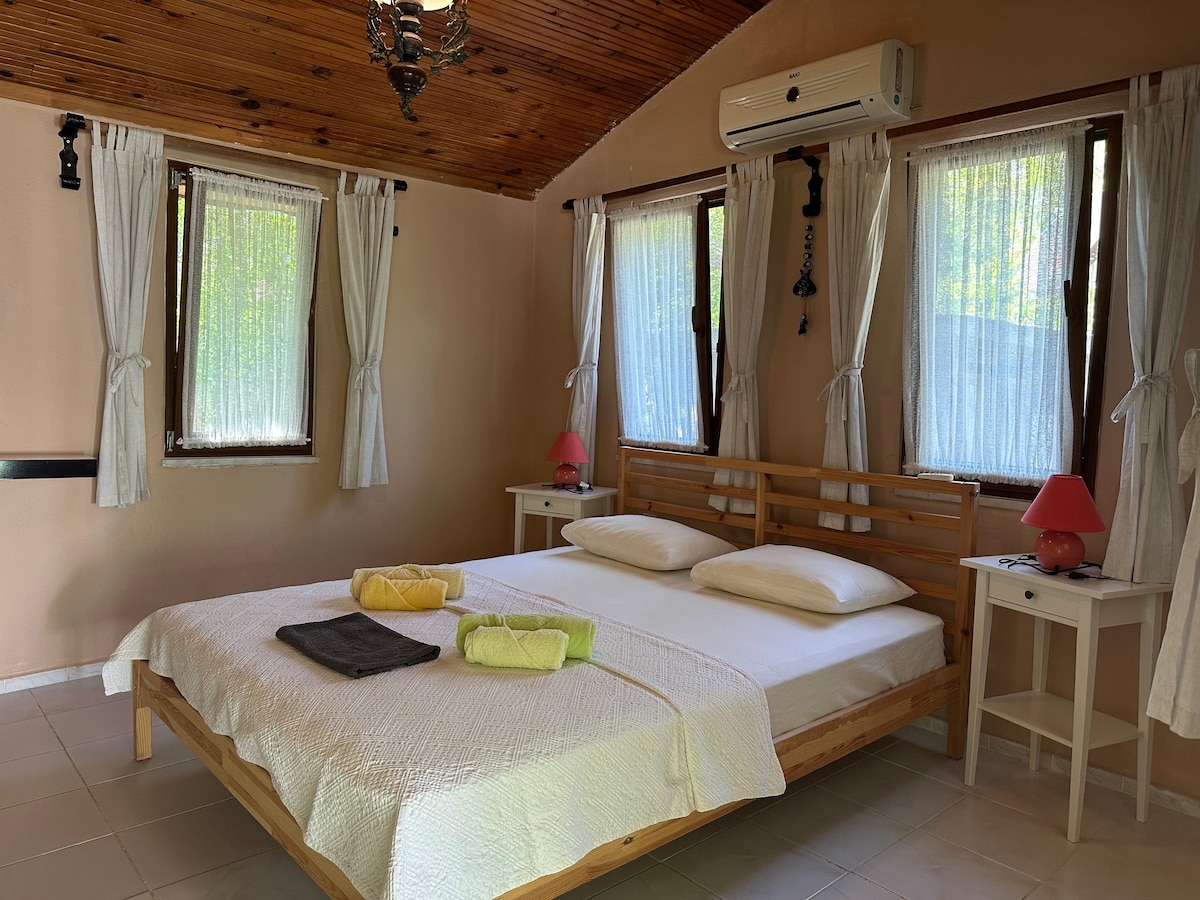 A spacious bedroom is furnished with a large wooden bed, topped with clean white linens. Two soft lamps in red shades sit on either side, while natural light filters through sheer, white curtains. Air conditioning is present for comfort.