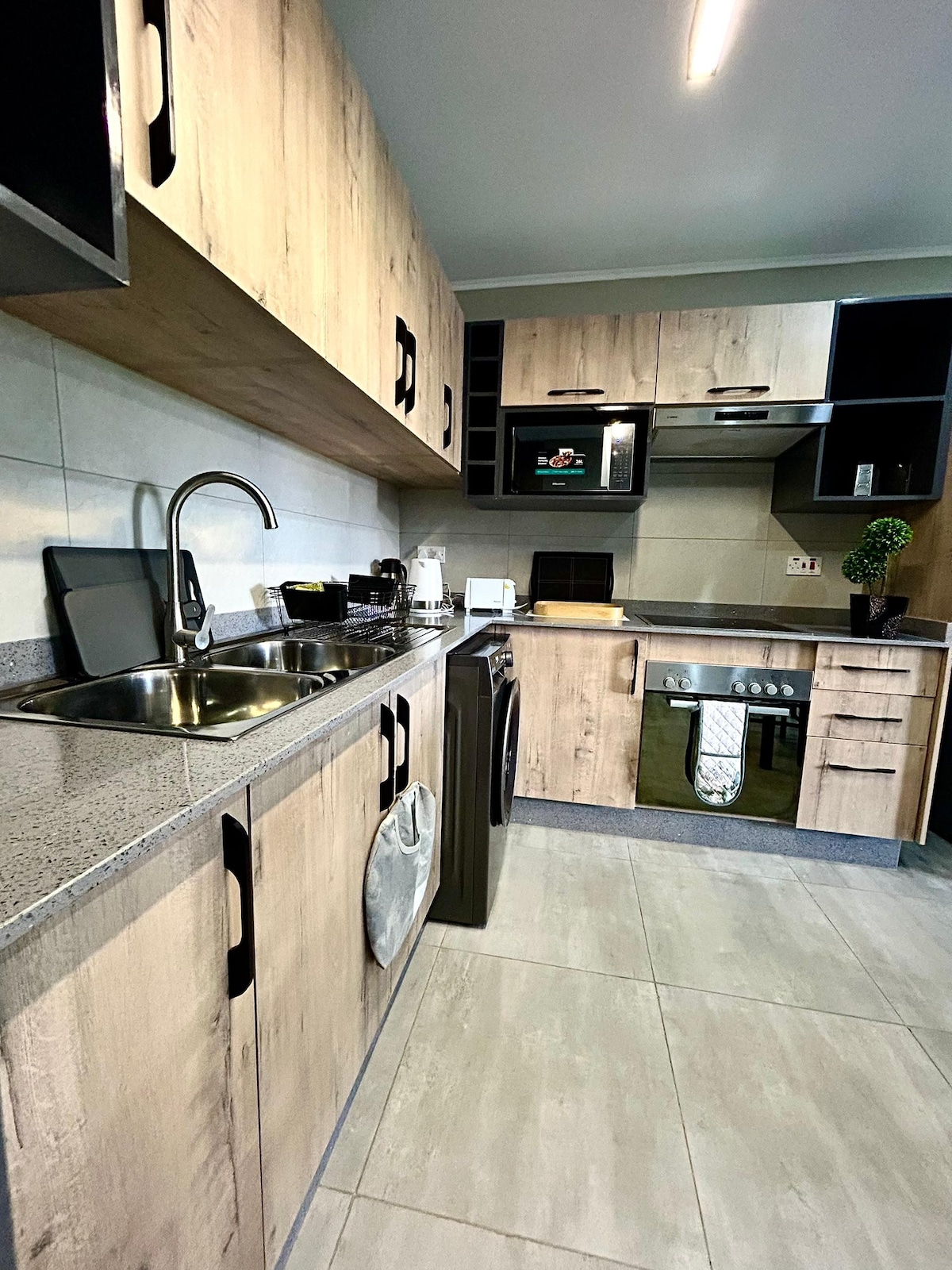 A modern kitchen features light wood cabinets and dark accents. Stainless steel appliances, including a microwave and washing machine, are arranged for efficiency. Counter space is provided for meal preparation, and a sink is positioned beneath a window with a small indoor plant for a touch of greenery.