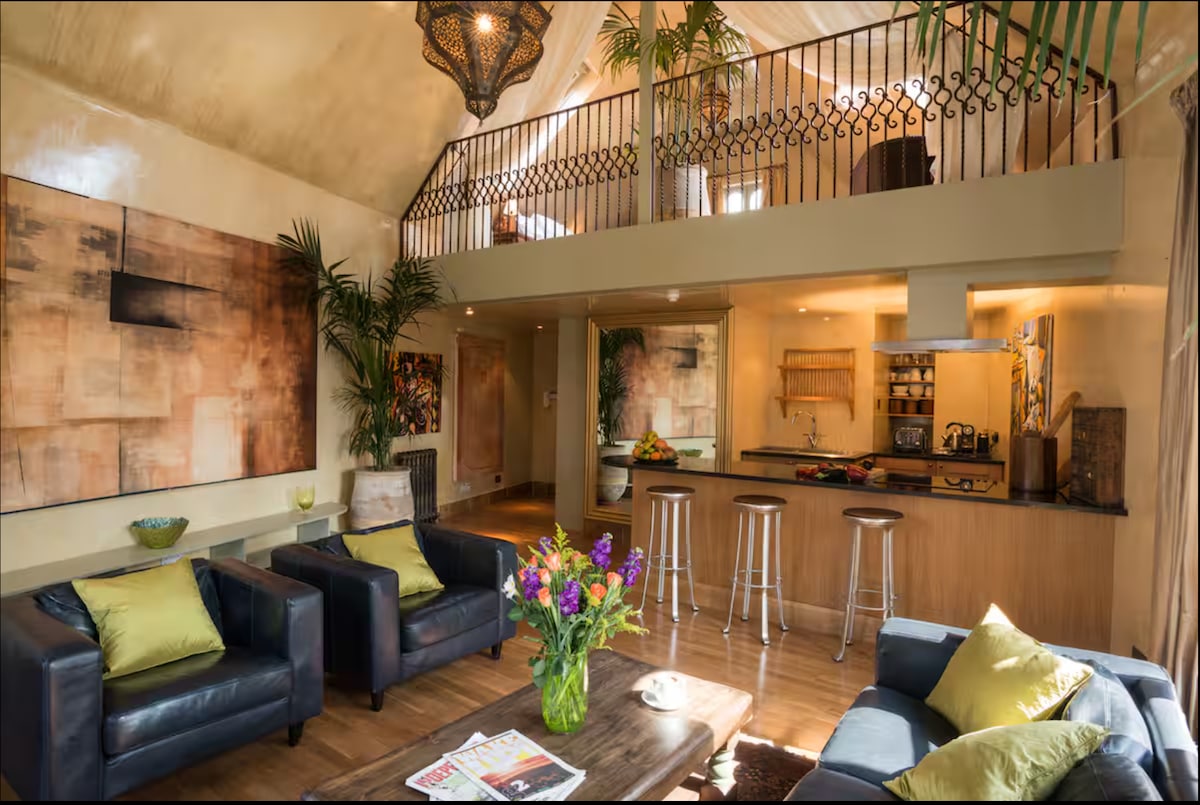 An open-plan living area features a relaxed seating arrangement with two dark chairs and a blue sofa adorned with green cushions. A coffee table sits centrally, complemented by a vibrant flower arrangement. The room is brightened by natural light streaming from the upper mezzanine, where a railing adds a decorative touch.