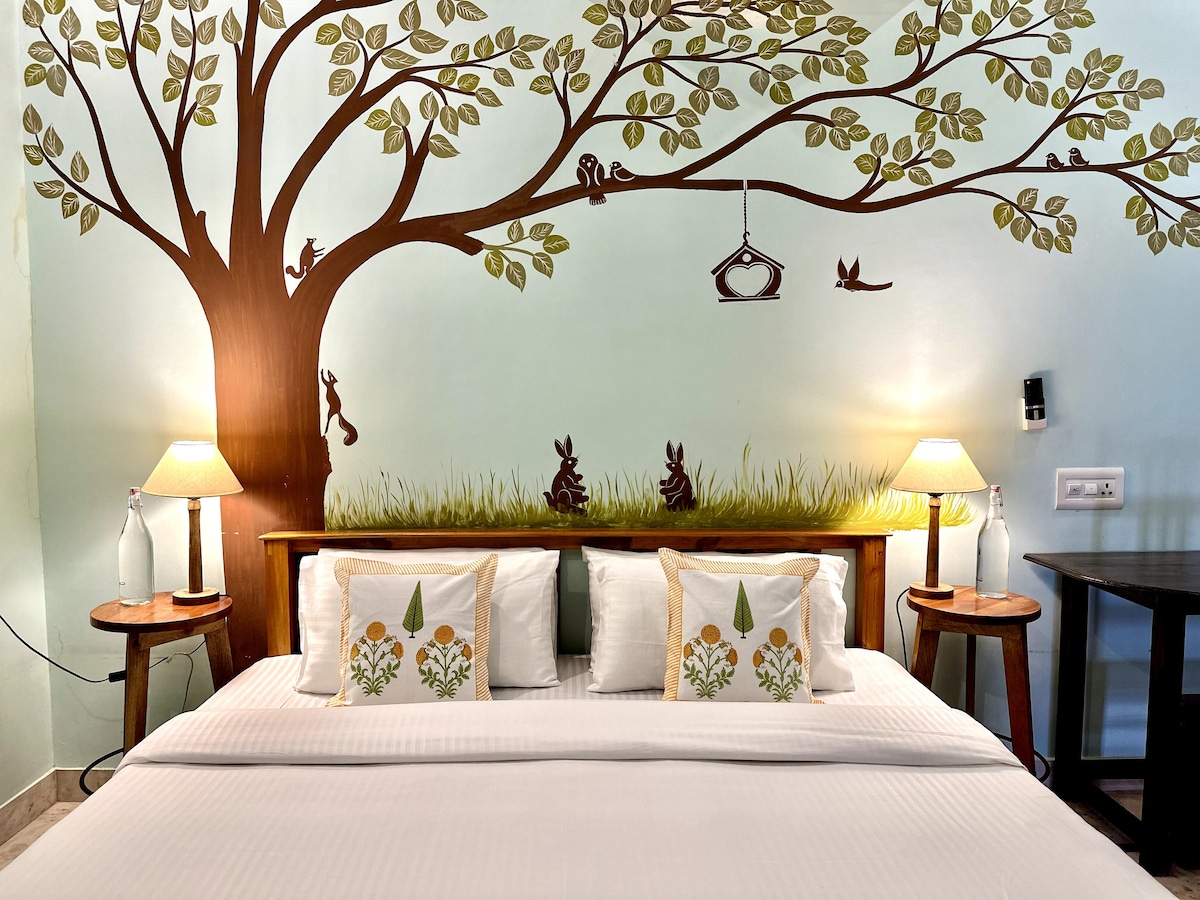 A serene sleeping area is highlighted by a decorative mural of a tree with playful animals. Two bedside lamps provide gentle lighting, and floral-patterned cushions adorn a spacious bed with crisp white linens. The walls are painted in soft blue, creating a calming atmosphere.