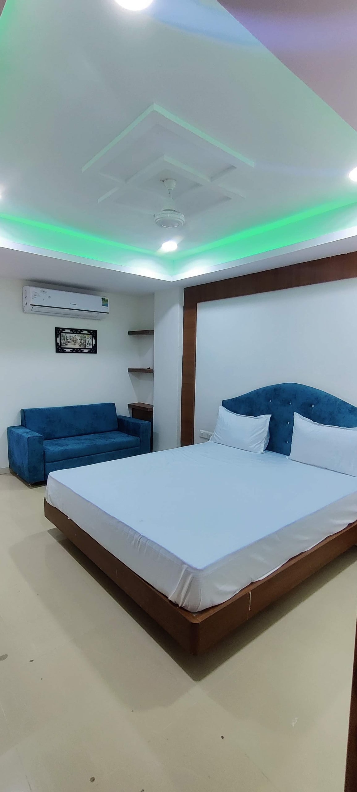 A modern room features a spacious bed with crisp white bedding. A small blue couch sits against the wall, complemented by a shelf nearby. Soft green lighting accents the ceiling, while a wall-mounted air conditioning unit offers comfort.
