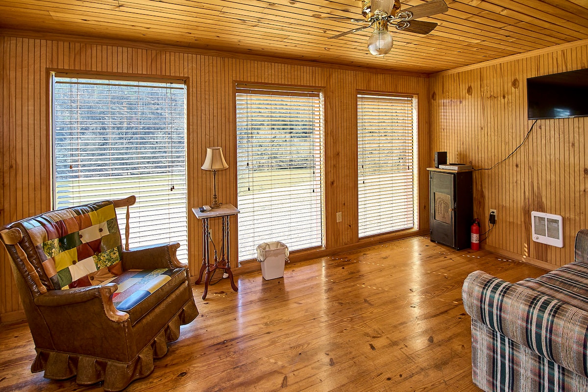 A spacious living area features wooden paneling and a ceiling fan, complemented by multiple windows that allow natural light to fill the room. A colorful patchwork sofa and a plaid chair provide comfortable seating. A small lamp and television add functional elements to the space.