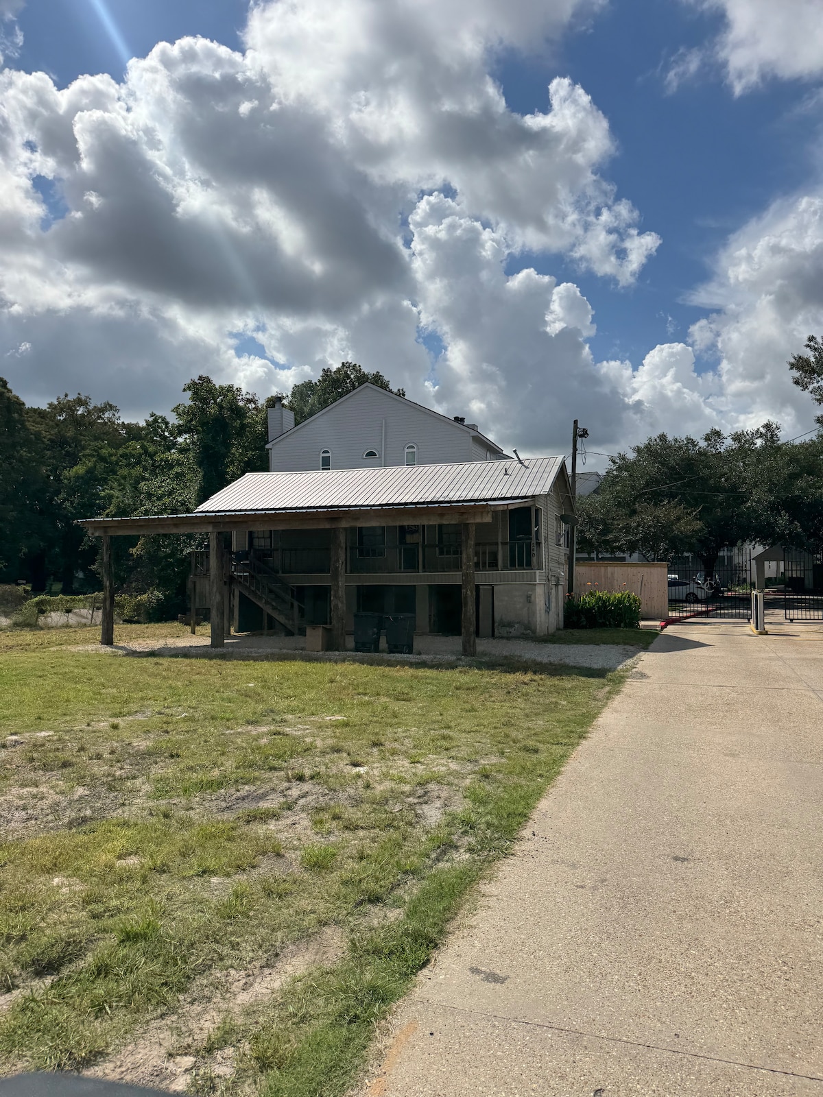 A two-story house is depicted, featuring a metal roof and wooden supports. The ground level is elevated, with a covered porch extending across the front. Surrounding greenery and open space contribute to the rural charm of the location. The backdrop includes a cloud-filled sky.