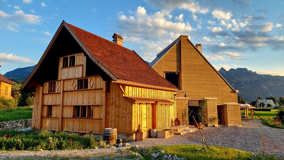 A historic wooden structure combines rustic charm with modern architecture, showcasing a warm wooden facade and a sloped roof. Lush green surroundings and a stone pathway lead to the entrance, with distant mountains visible under a sky filled with soft, scattered clouds.
