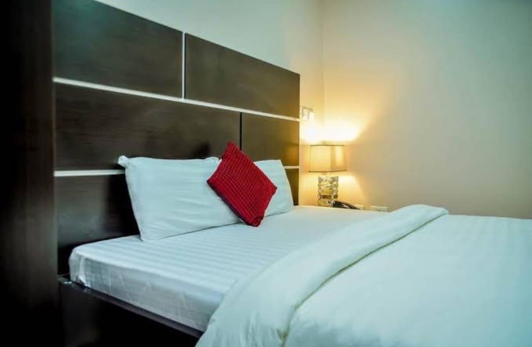 A spacious bed is positioned against a dark wooden headboard. A red decorative pillow adds a pop of color. Soft lighting from a bedside lamp casts a warm glow on the neutral walls, creating a calming atmosphere.