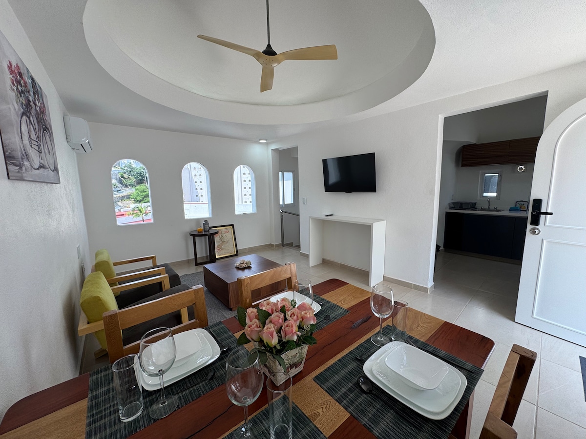An open living space features a circular ceiling with a ceiling fan. A wooden dining table is set for multiple guests, surrounded by glasses and plates. Comfortable seating is arranged across from a wall-mounted television, with arched windows allowing natural light to fill the area.