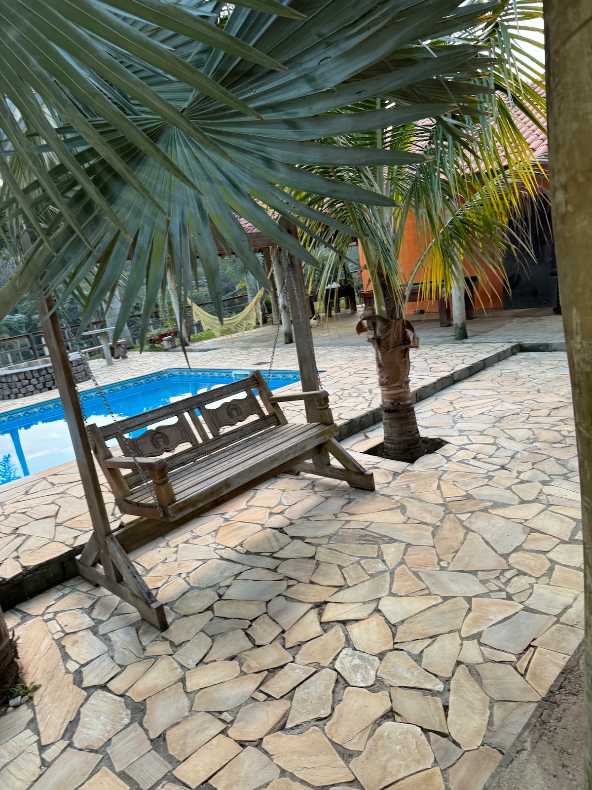 A swing bench is positioned on a stone patio, surrounded by tropical palm trees. A swimming pool is visible in the background, its clear water reflecting the nearby greenery. A warm color from a structure is seen providing an inviting ambiance.