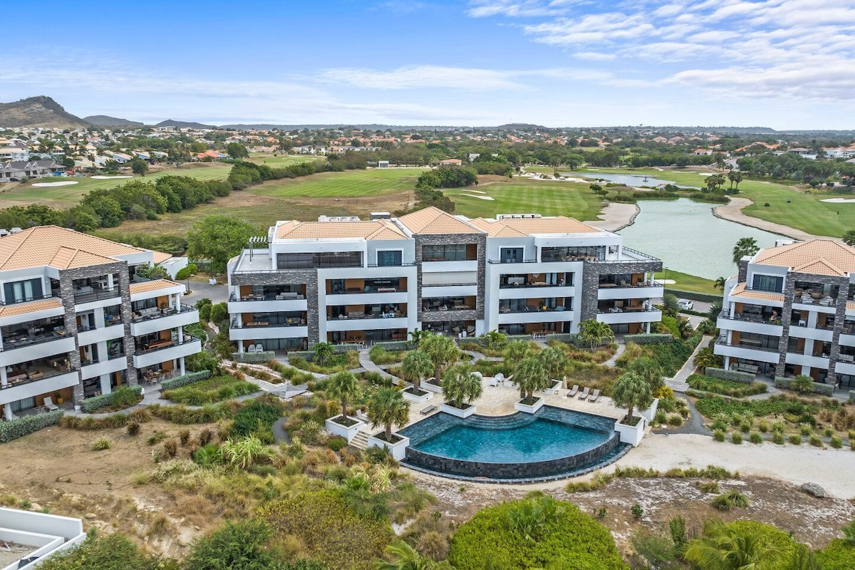 An aerial view of a modern apartment complex is presented, highlighting the lush landscaping and private pool area. The building is framed by greenery and overlooks a golf course, with clear skies in the background.