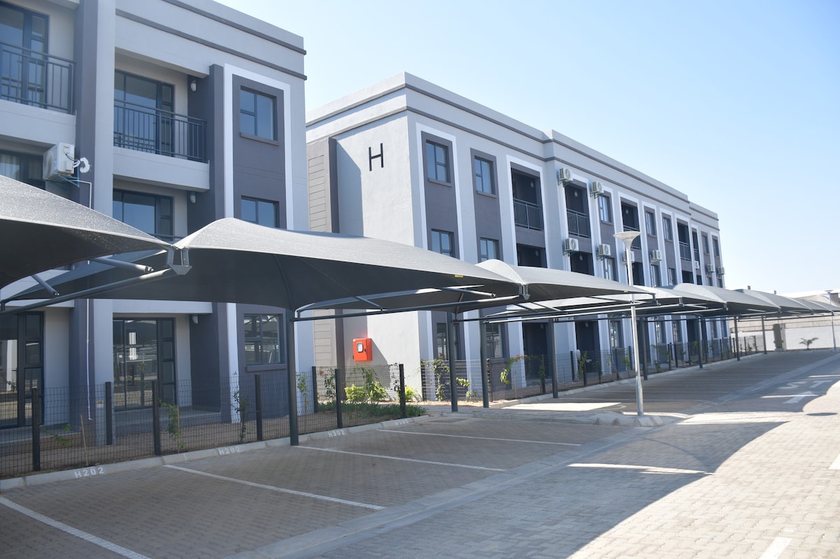 A modern building features multiple stories with a sleek design and balconies on the upper levels. Covered parking spaces are visible in the foreground, with a neatly paved area and greenery along the perimeter, providing a tidy and organized appearance.