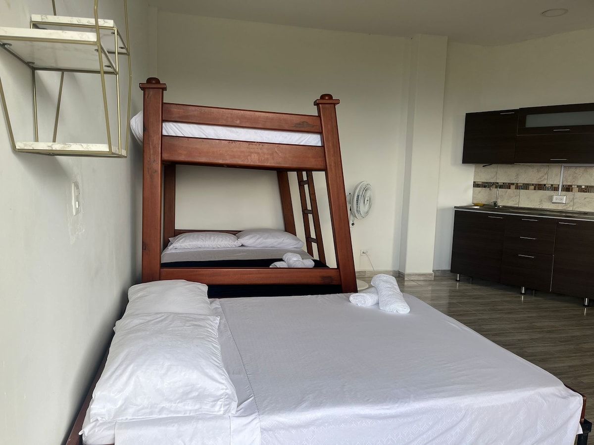 A spacious room features a double bed on the lower level and a bunk bed above. Light-colored linens are neatly arranged on both beds. A simple shelving unit is mounted on the wall, and a kitchenette is visible in the background, equipped with dark cabinetry.
