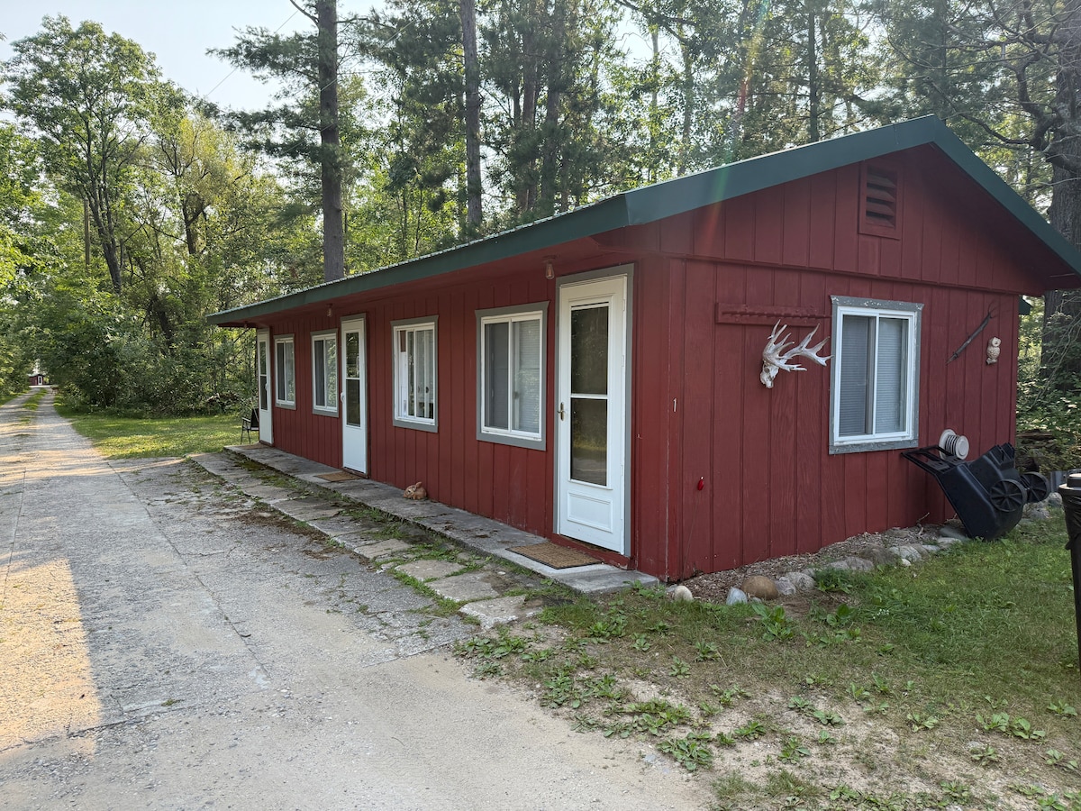 The exterior of a red cabin is shown, situated among trees. A gravel path runs alongside the structure, which features multiple windows and a front door. An antler decorates the wall, and outdoor items are positioned nearby.