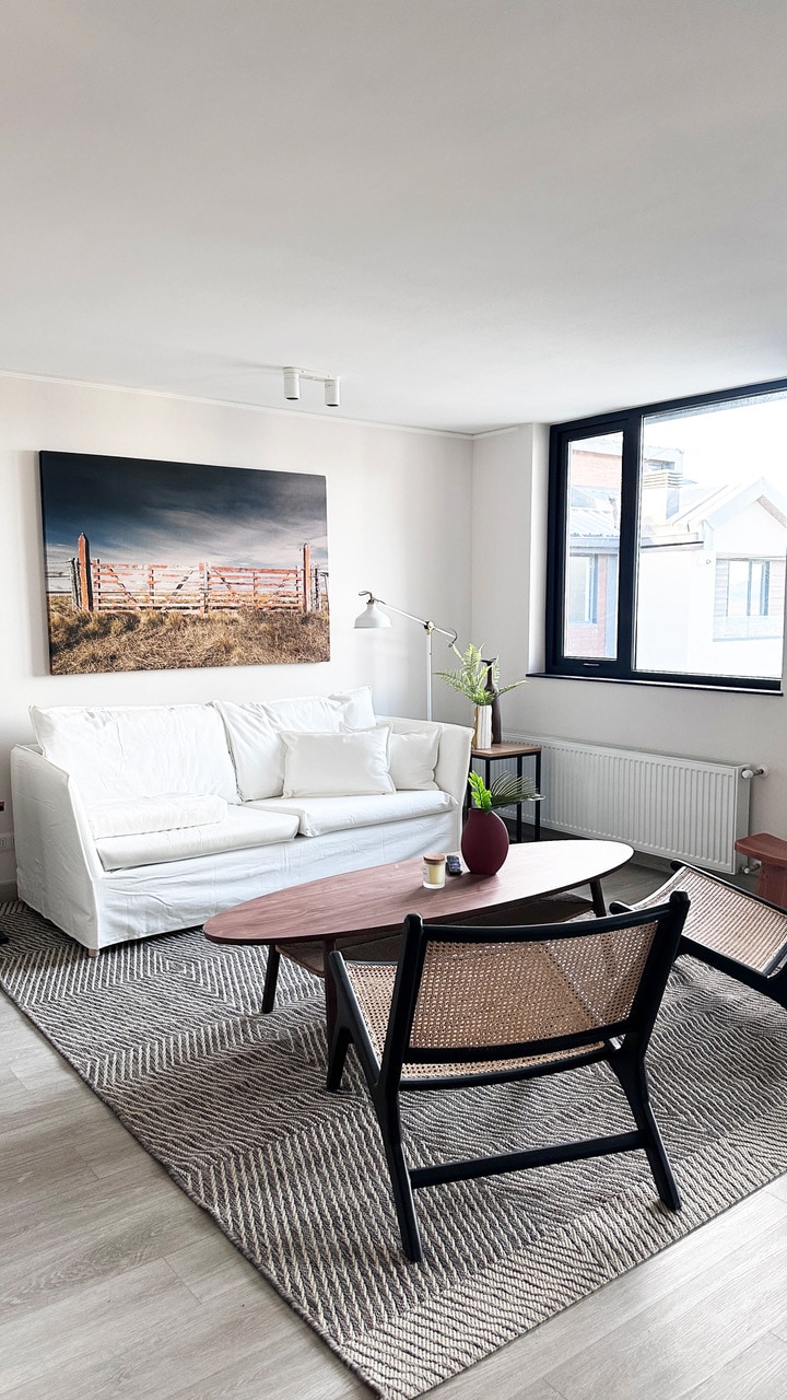 A light-filled living area contains a white sofa positioned beside a wooden coffee table. A woven rug covers the floor. A floor lamp and a vase with greenery complement the space, while a large artwork depicting a fence landscape adorns the wall. Large windows provide natural light.