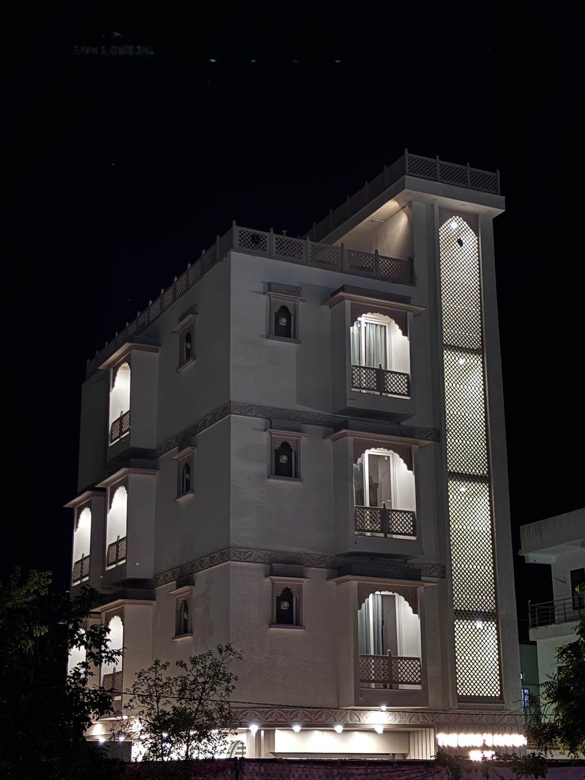 A multi-story building features a white facade with intricate architectural details and decorative balcony railings. Soft lighting illuminates the exterior, highlighting the windows and design elements. Trees frame the structure, adding a touch of greenery to the nighttime scene.