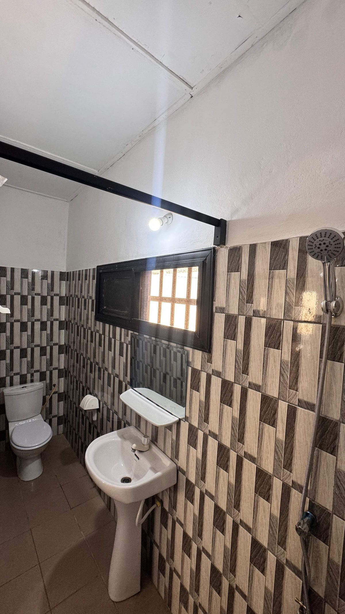A modern bathroom features tiled walls with a mix of light and dark patterns. A white toilet is positioned next to a sink, with a mirror mounted above it. A showerhead is installed on the far wall, and natural light enters through a small window.