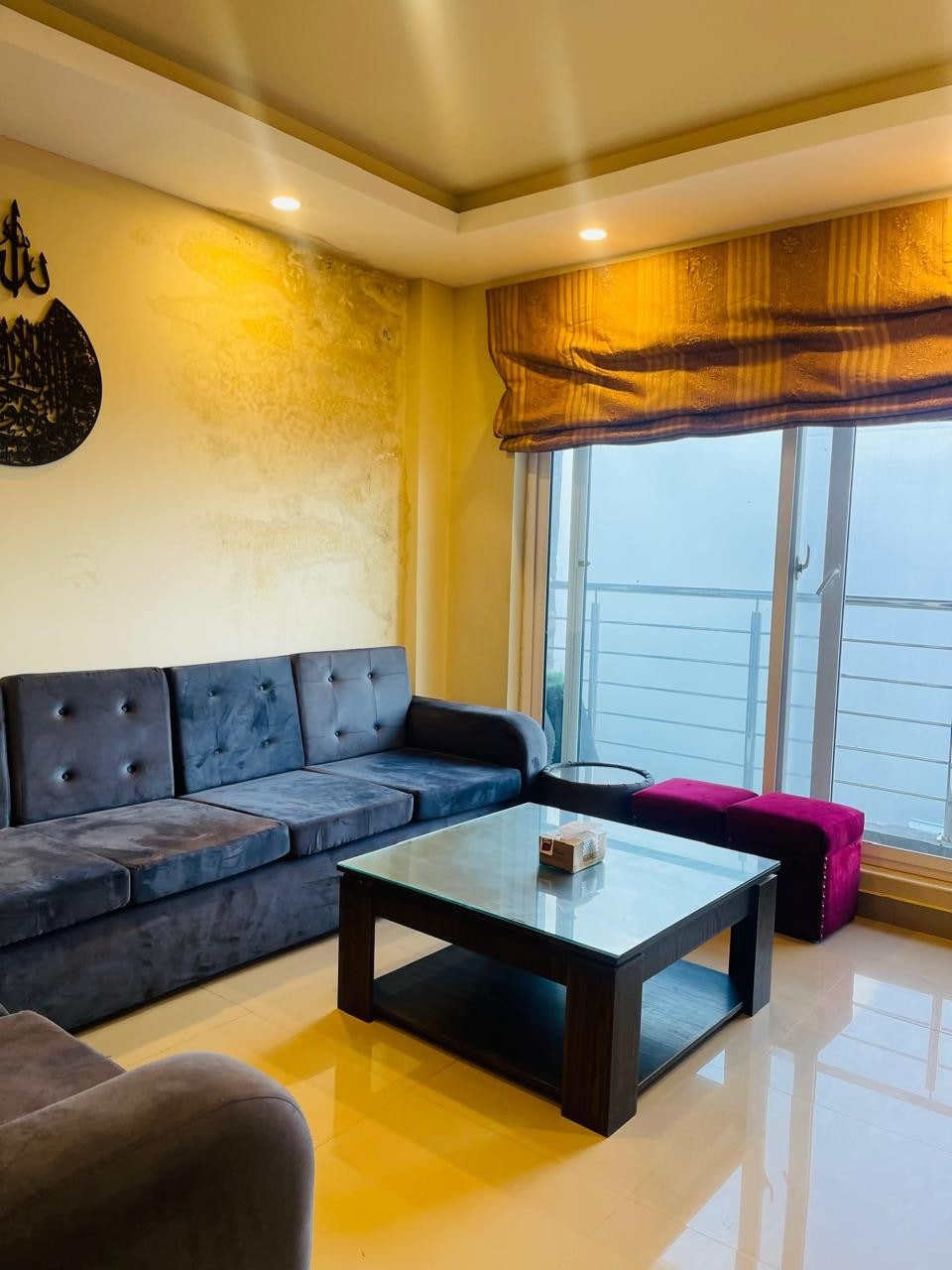A cozy living area features a comfortable dark-gray sofa and a plush golden window treatment allowing soft light to enter. A wooden coffee table sits in the center, accompanied by a vibrant pink ottoman. A large window offers a view of the serene surroundings.