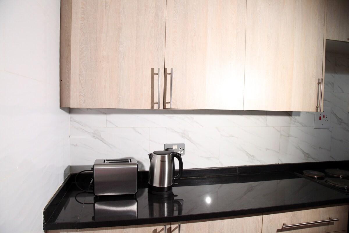 A sleek kitchen space features light wooden cabinetry and a polished black countertop. A stainless steel toaster and an electric kettle are displayed on the counter, complemented by a minimalist backsplash. The setting conveys a modern aesthetic suitable for meal preparation.