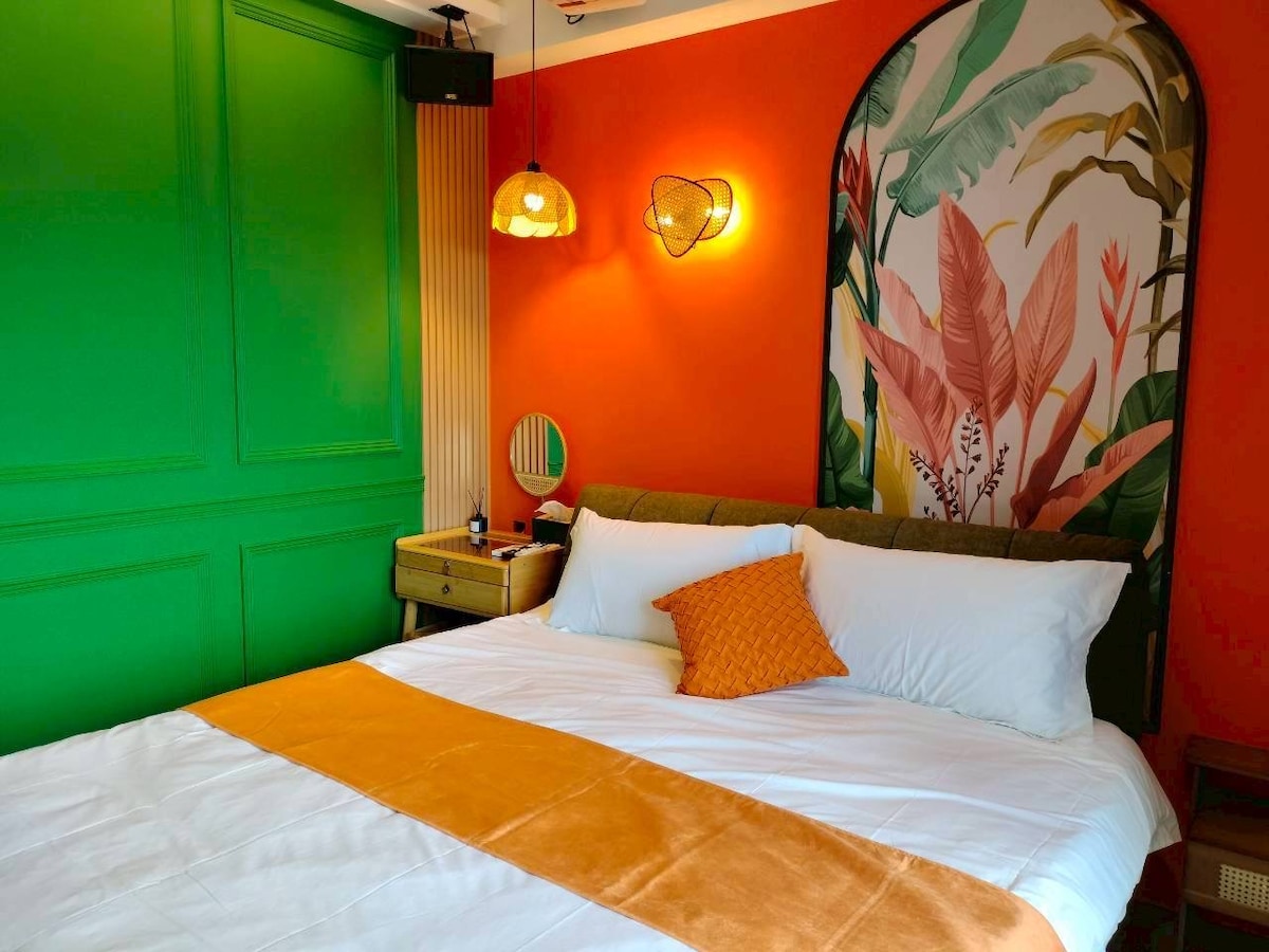 A vibrant sleeping area features a spacious bed with crisp white linens and an orange accent pillow. The walls are adorned in a striking green and warm orange, complemented by decorative lighting and a botanical wall art piece, creating a lively and welcoming ambiance.