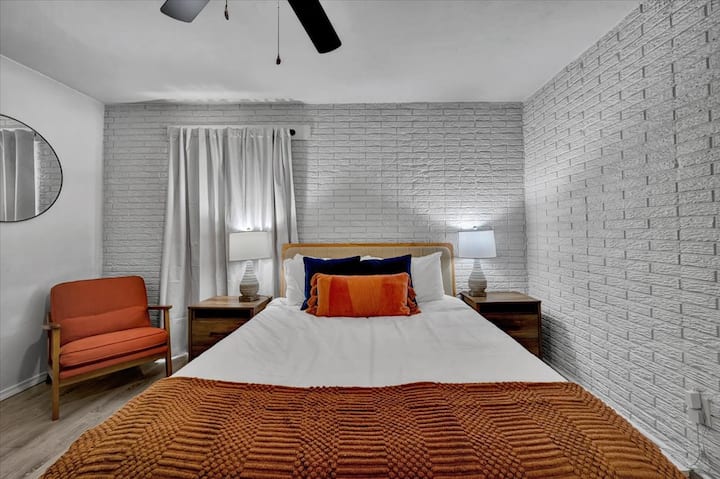 Moderno uređena spavaća soba s bijelim zidom od cigle, toplim narančastim i plavim tonovima te simetričnim noćnim svjetiljkama. Ima udobnu fotelju, stropni ventilator i složenu posteljinu – savršeno za mirne noći u pažljivo uređenom prostoru.