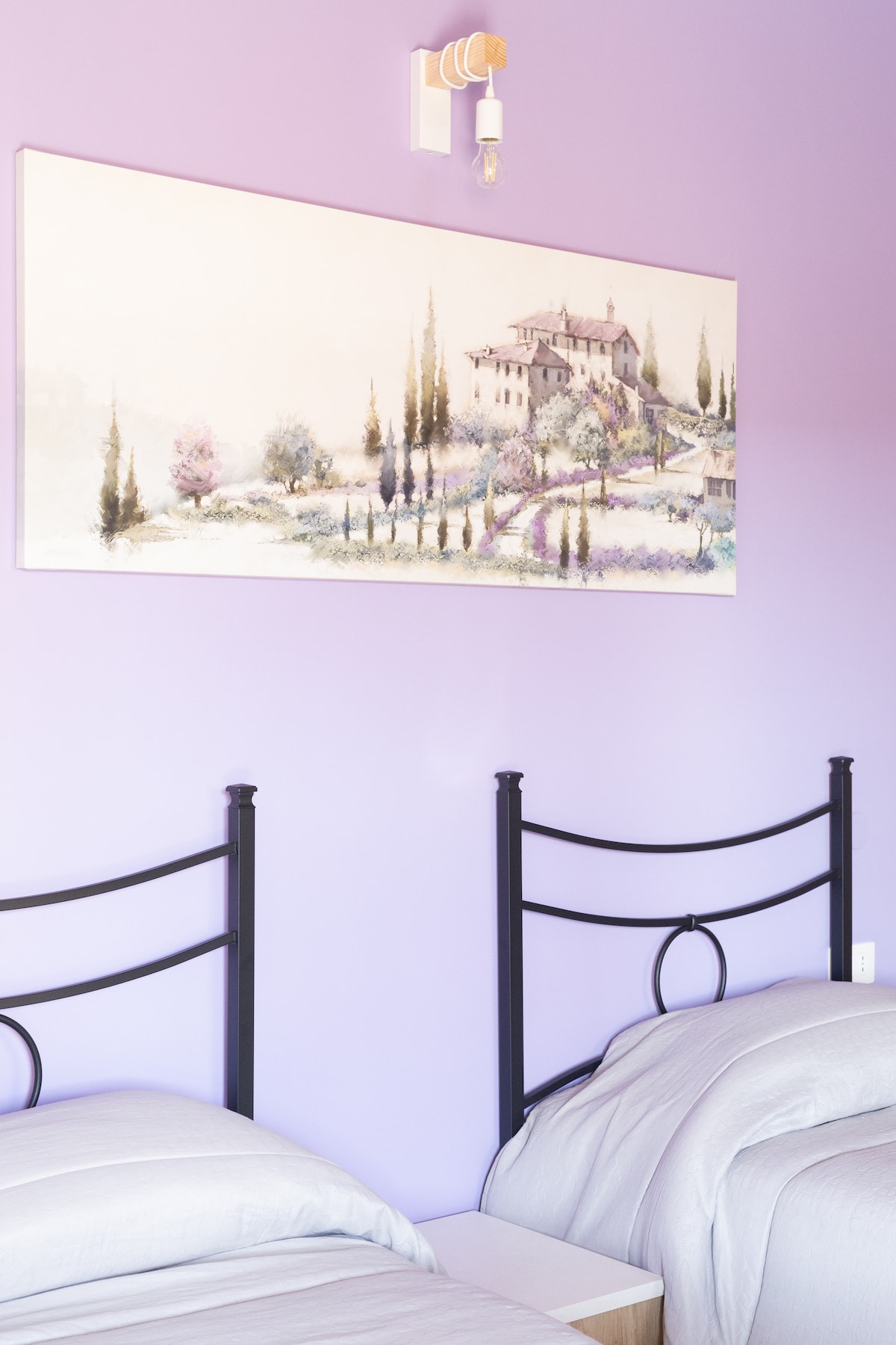 A serene sleeping area features two beds with light grey linens, positioned against a soothing purple wall. Above, a landscape painting depicts a gentle hillside view, adding a calming touch to the space. A wooden nightstand is placed between the beds.
