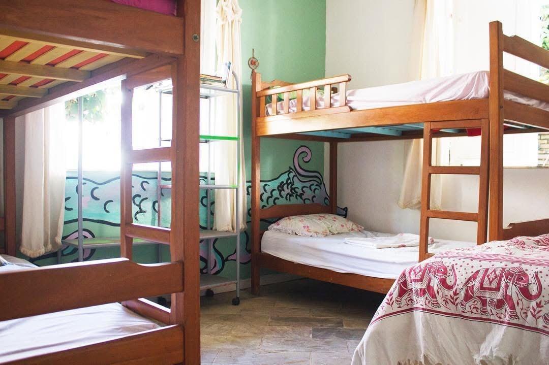 A bright shared sleeping area features two wooden bunk beds and a single bed. Walls are painted in soft green with a colorful mural depicted at the base. Natural light filters through partially open curtains, creating a relaxed atmosphere.