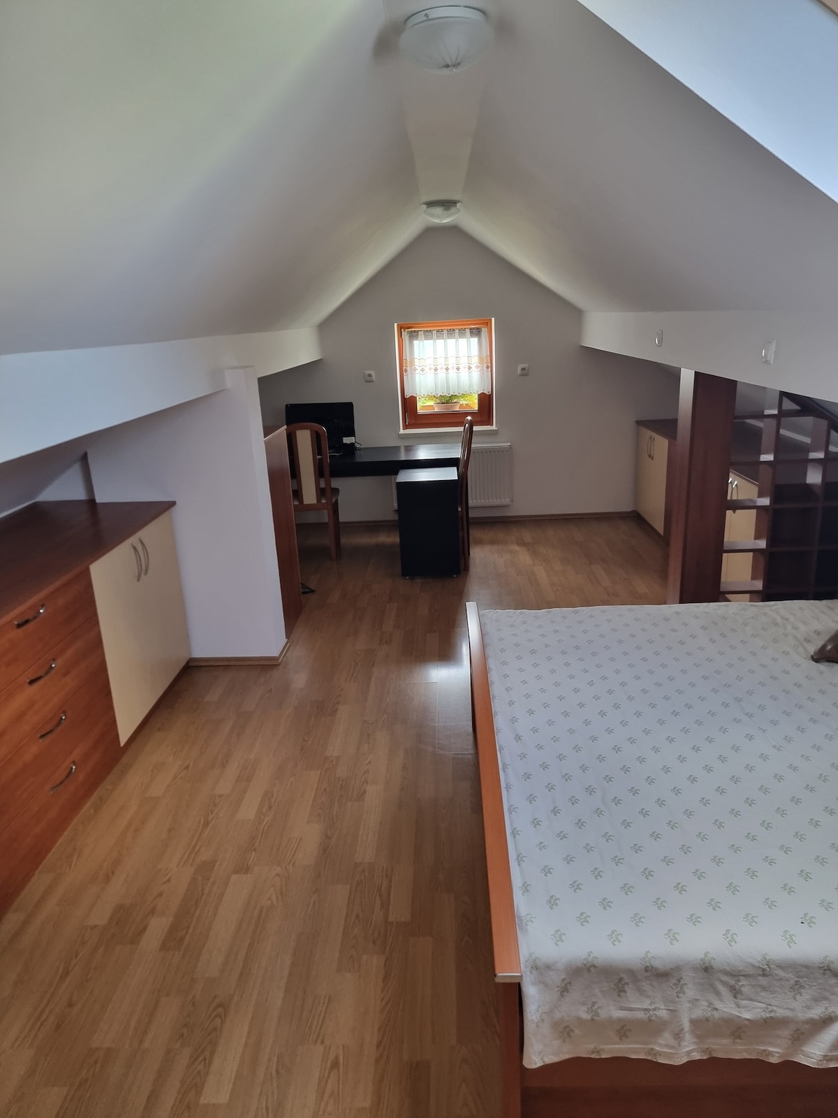 An airy attic room features a spacious bed alongside a work desk and chair. Light wood flooring extends throughout the area, complemented by built-in storage units against the walls. A ladder leads to a lofted space, enhancing the room's vertical feel.