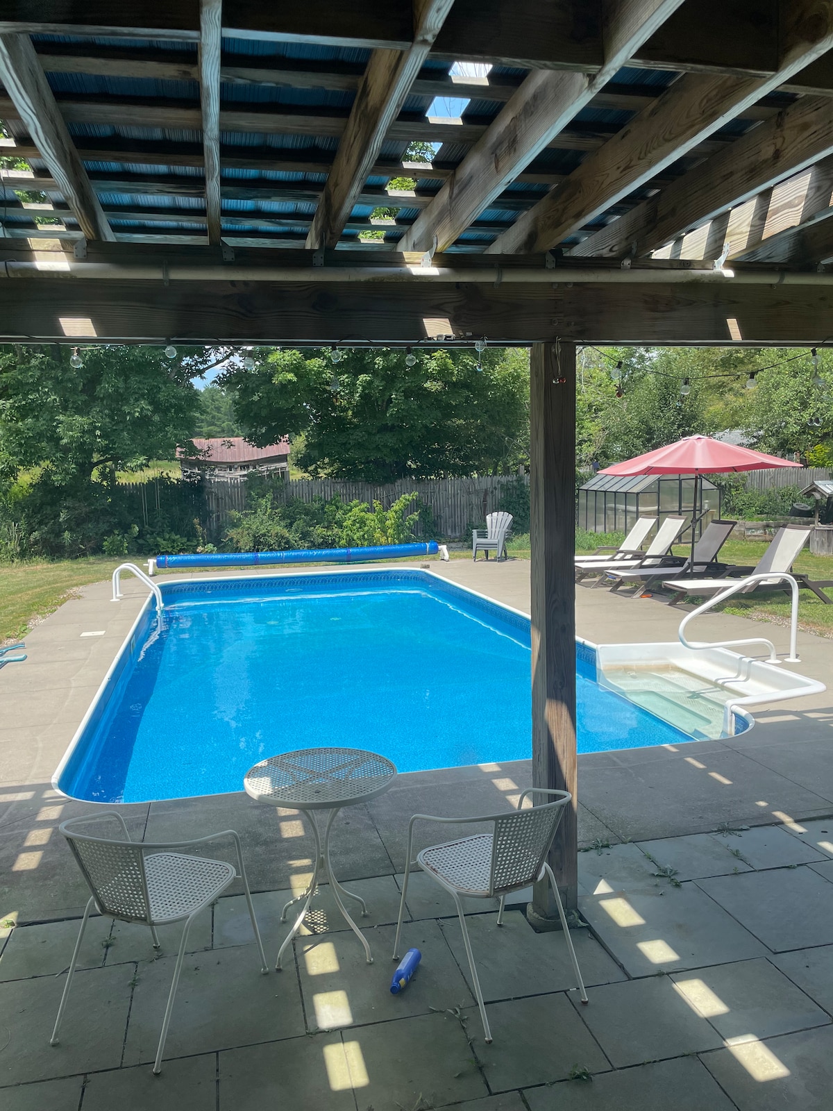 A pool area is framed by a wooden overhang, showcasing a clear blue swimming pool surrounded by a patio. Lounge chairs are neatly positioned nearby, and an umbrella provides shade. Lush greenery and a privacy fence create a serene outdoor environment.