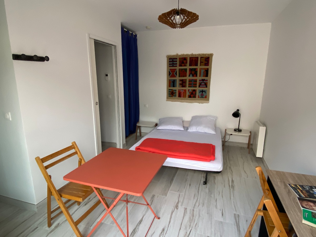 A cozy studio space features a spacious bed adorned with white linens and a vibrant orange throw. A wooden table with two folding chairs stands nearby. A colorful wall art piece adds a touch of character, while blue curtains frame a window that brings in natural light.