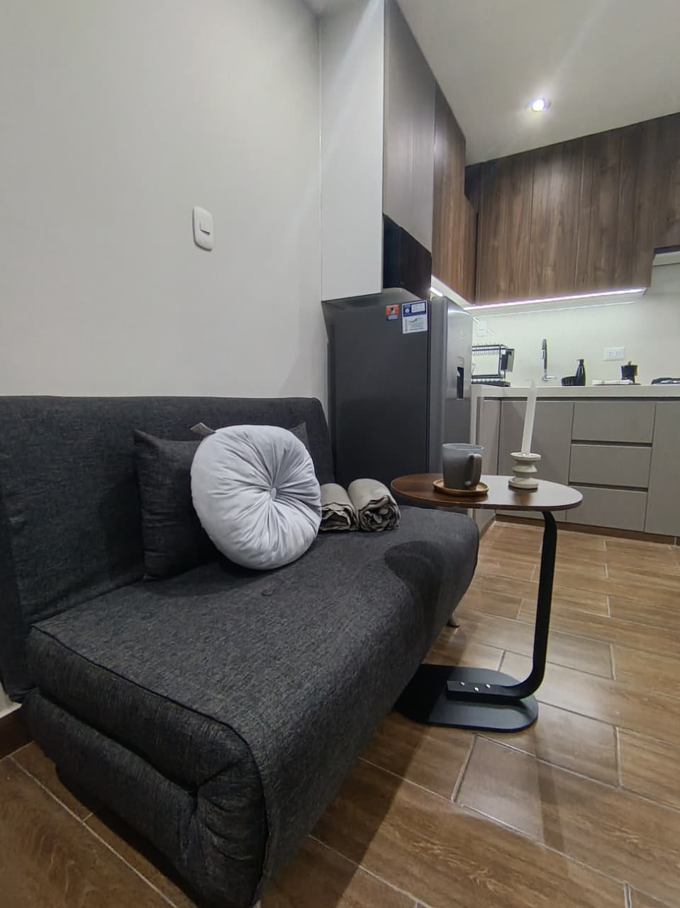 A modern sitting area features a dark gray sofa adorned with a decorative pillow and cozy throws. A small round table holds a cup and candle, while integrated cabinetry and appliances are visible in the background, highlighting a functional kitchen space.