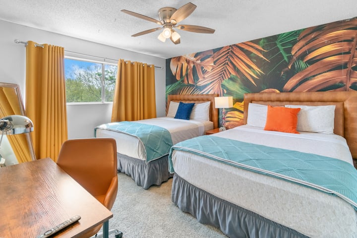 Dormitorio 2 con 2 camas queen, mural de palmeras tropicales, televisión inteligente y luz natural; capacidad para 4 personas cómodamente.