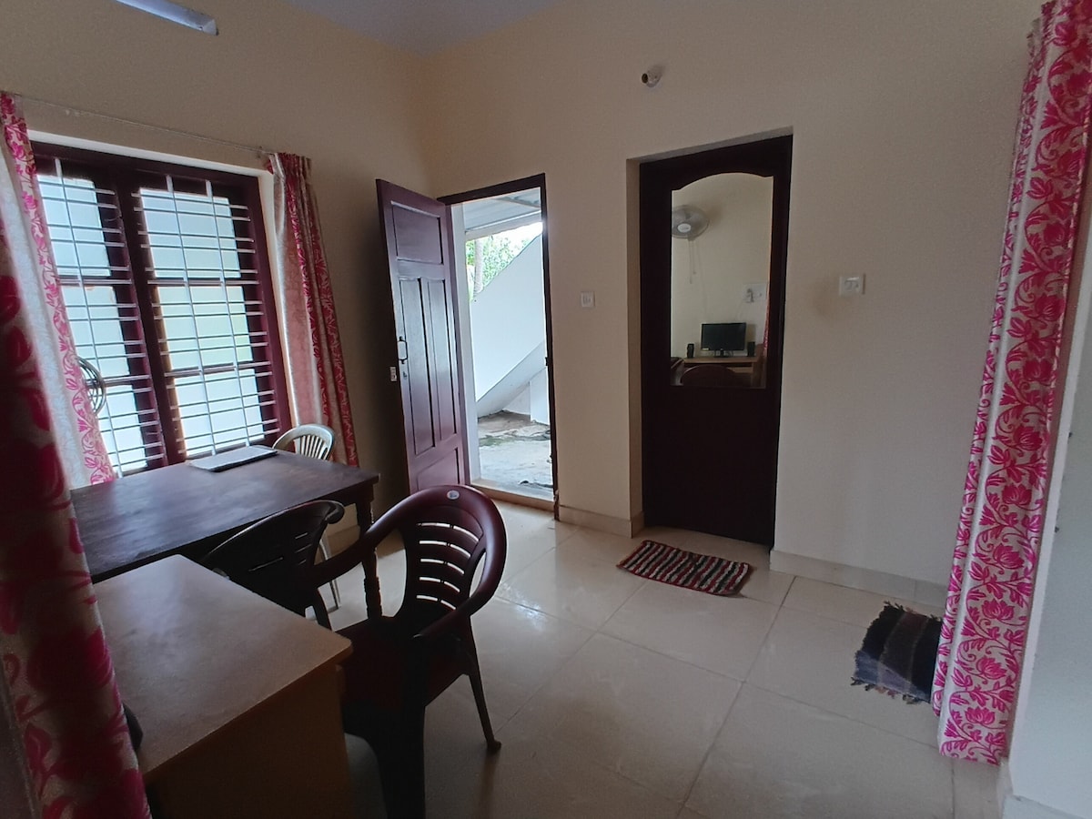 A well-lit interior space is highlighted, featuring a dining table accompanied by a pair of dark chairs. A doorway connects to an outdoor area, while a second door leads to another room. Light-colored walls and tiled flooring create an open and inviting atmosphere.