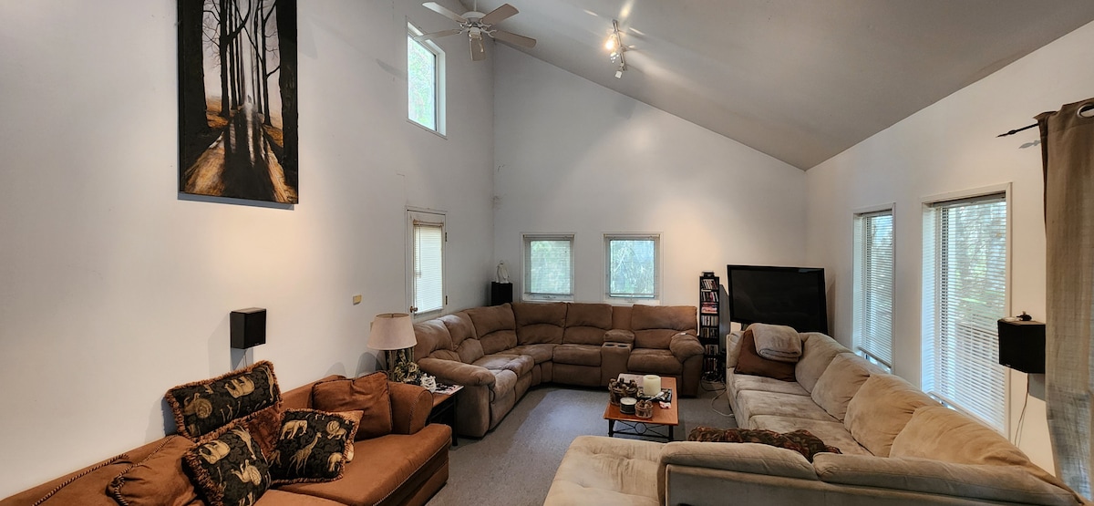 A spacious living area features two large sectional couches in neutral tones, arranged to encourage social gatherings. Tall windows allow natural light to fill the room. A television and art piece adorn the wall, complementing the inviting atmosphere of the space.