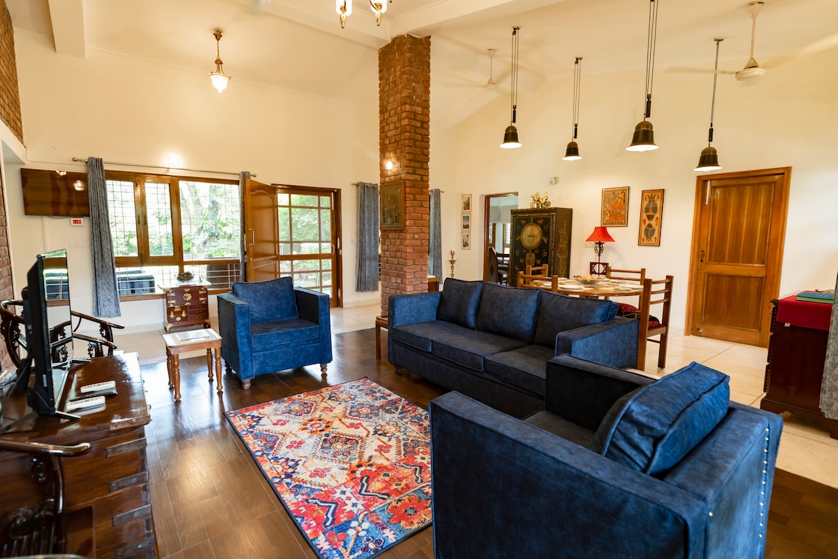 The spacious living room features dark blue sofas and armchairs, arranged on a vibrant area rug. Natural light fills the space through large windows, highlighting the brick wall details. A wooden dining table and decorative elements complete the inviting atmosphere for gatherings.